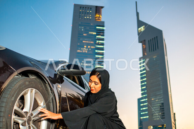 Women's self-reliance in movement and transportation, the concept of independence and self-reliance, a veiled Emirati Arab woman sitting next to her car, freedom of driving for Arab women, a Saudi woman wearing an abaya checking the car's tires, the backg