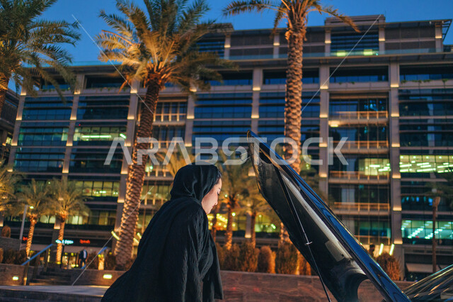 The concept of independence, freedom and self-reliance, a veiled Emirati Arab Gulf woman standing near her broken car and opening the hood, freedom of driving for Arab women, a side image of a Saudi woman wearing an abaya repairing and checking the car, t