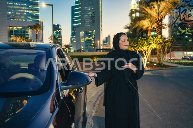 حركة يدين تدل على الاشارة لداخل السياره، حرية القيادة للمرأة العربية، سيده سعودية تشير بيديها الى السيارة، اعتماد المرأه على نفسها في التنقل و المواصلات, مفهوم الاستقلال و الحرية و الاعتماد على النفس، امرأه عربية خليجية اماراتيه محجبة ترتدي العباءة تقف بج