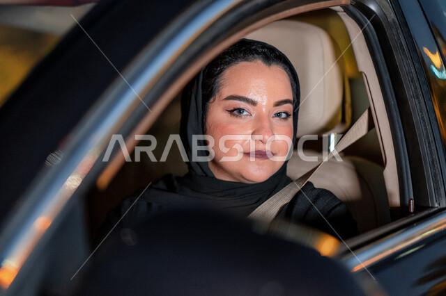 سيدة إماراتيه محجبة تجلس داخل سيارتها تنظر لشيء ما، اتباع قوانين الحركة المرورية، إتقان قيادة المرأة للسيارة في الطرق الرئيسية، إمرأة عربية سعودية خليجية ترتدي حزام الأمان تقود بحذر، مفهوم الاستقلال والاعتماد الذاتي، الإلتزام بقواعد الأمن والسلامة العامة.