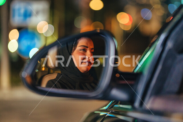 القيادة بحذر واتباع قواعد الأمن والسلامة العامة، شابة سعودية محجبة تجلس داخل سيارتها تنظر لشيء ما، صورة مقربة لانعكاس وجه سيده عربية اماراتيه خليجية مبتسمة على مرآة السيارة الجانبية، اتقان المرأة لقيادة السيارة في الطرق العامة، مفهوم الحرية والاستقلال.