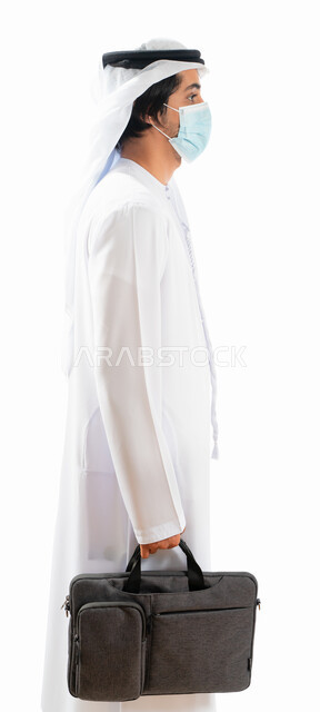 Portrait from the side of an Arab Gulf Emirati businessman wearing a protective mask and carrying a work bag, standing straight, going to work, adhering to the measures to prevent corona virus, white background