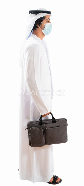 Portrait of an Arab Gulf Emirati businessman wearing a protective muzzle and carrying a work bag, moving forward, gestures indicating movement and walking, going to work, adherence to measures to prevent corona virus, full-length body image, white backgro