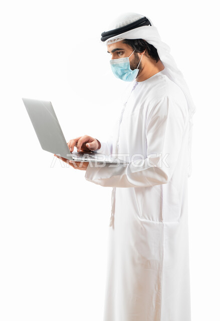 بورتريه لرجل أعمال عربي خليجي اماراتي يرتدي الكمامة الواقية، انجاز مهام العمل من خلال الحاسوب المحمول، العمل عن بعد، مفهوم إدارة الأعمال، بيئة عمل، الالتزام باجراءات الوقاية من فايروس كورونا، اعمال مكتبية، خلفية بيضاء