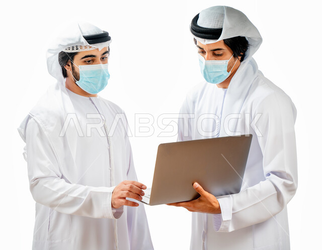 Portrait of two Arab Gulf Emirati businessmen wearing a protective muzzle, completing work tasks through a laptop computer, business discussions and dialogues, work follow-up, Emirati work environment, supervision and work auditing, adherence to coronavir
