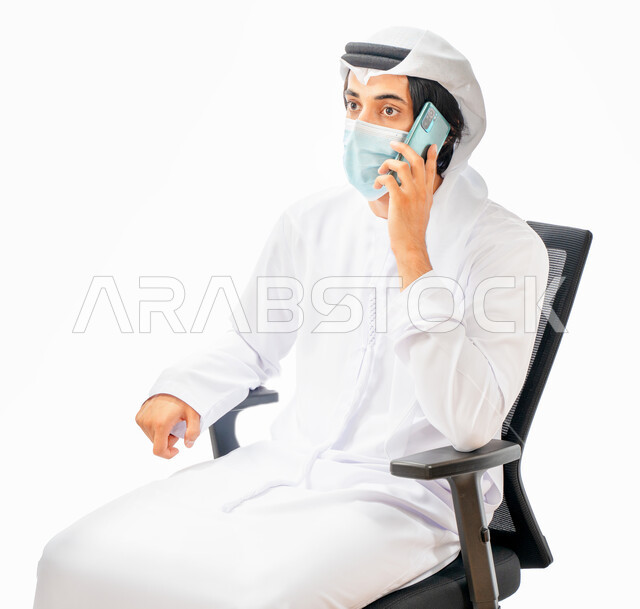 Portrait of an Emirati Arab Gulf businessman wearing a protective muzzle and making a phone call, sitting on a chair, calling and chatting, communicating with customers, making deals, working remotely, e-marketing, adherence to measures to prevent corona 