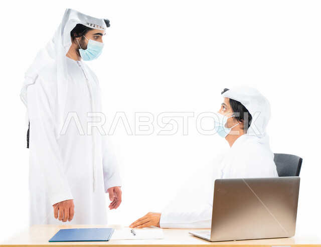Portrait of two Arab Gulf Emirati businessmen wearing a protective muzzle, completing work tasks through a laptop computer, business discussions and dialogues, work follow-up, Emirati work environment, supervision and work auditing, adherence to coronavir