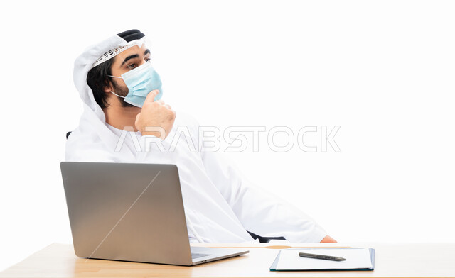 Portrait of an Emirati Arab Gulf businessman wearing a protective muzzle, completing work tasks through a laptop, remote work, Emirati work environment, adherence to coronavirus prevention measures, gestures and expressions indicating thinking and confusi