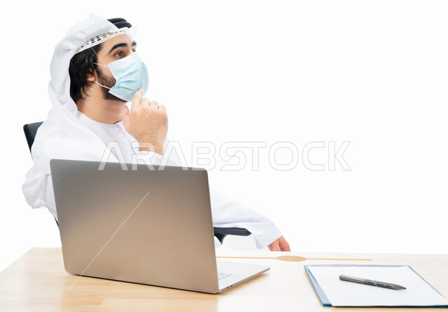 Portrait of an Emirati Arab Gulf businessman wearing a protective muzzle, completing work tasks through a laptop, remote work, Emirati work environment, adherence to coronavirus prevention measures, gestures and expressions indicating thinking and confusi