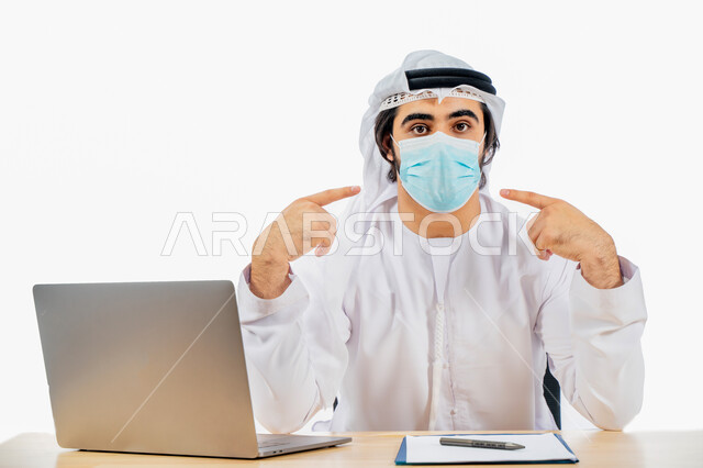 Portrait of an Arab Gulf Emirati businessman wearing a protective muzzle and pointing his fingers towards the muzzle, completing work tasks through a laptop computer, remote work, business management concept, work environment, adherence to coronavirus pre