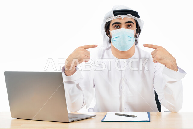 بورتريه لرجل أعمال عربي خليجي اماراتي يرتدي الكمامة الواقية ويشير باصبعيه نحو الكمامة، انجاز مهام العمل من خلال الحاسوب المحمول، العمل عن بعد، مفهوم إدارة الأعمال، بيئة عمل، الالتزام باجراءات الوقاية من فايروس كورونا، اعمال مكتبية، خلفية بيضاء
