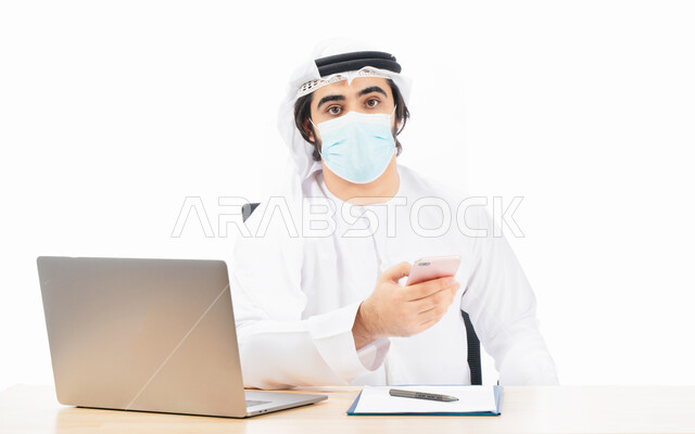 بورتريه لرجل أعمال عربي خليجي اماراتي يرتدي الكمامة الواقية ويستخدم الهاتف المحمول، انجاز مهام العمل من خلال الحاسوب المحمول، العمل عن بعد، مفهوم إدارة الأعمال، بيئة عمل، الالتزام باجراءات الوقاية من فايروس كورونا، اعمال مكتبية، خلفية بيضاء