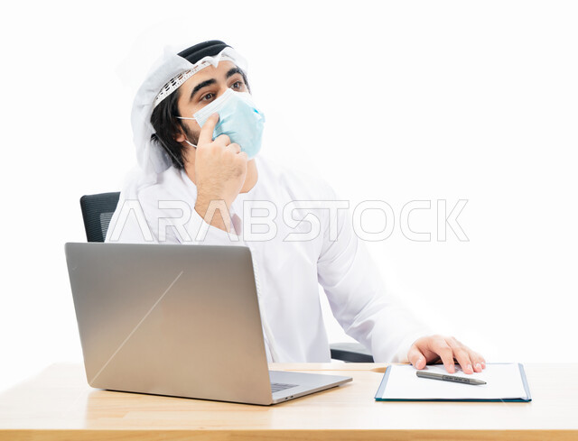 بورتريه لرجل أعمال عربي خليجي اماراتي يرتدي الكمامة الواقية، انجاز مهام العمل من خلال الحاسوب المحمول، العمل عن بعد، بيئة العمل الاماراتي، الالتزام باجراءات الوقاية من فايروس كورونا، ايماءات وتعابير تدل على التفكير والحيرة، اعمال مكتبية، خلفية بيضاء