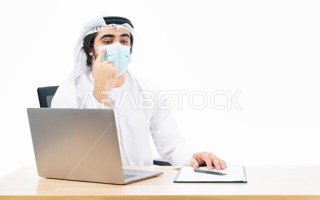 Portrait of an Emirati Arab Gulf businessman wearing a protective muzzle, completing work tasks through a laptop, remote work, Emirati work environment, adherence to coronavirus prevention measures, gestures and expressions indicating thinking and confusi