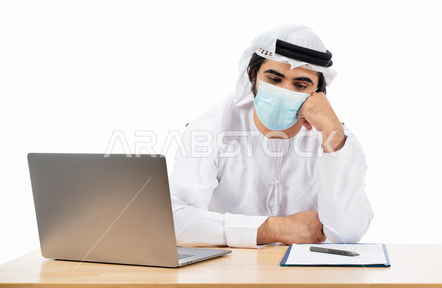 بورتريه لرجل أعمال عربي خليجي اماراتي يرتدي الكمامة الواقية، انجاز مهام العمل من خلال الحاسوب المحمول، العمل عن بعد، بيئة العمل الاماراتي، الالتزام باجراءات الوقاية من فايروس كورونا، ايماءات وتعابير تدل على التفكير والحيرة، اعمال مكتبية، خلفية بيضاء