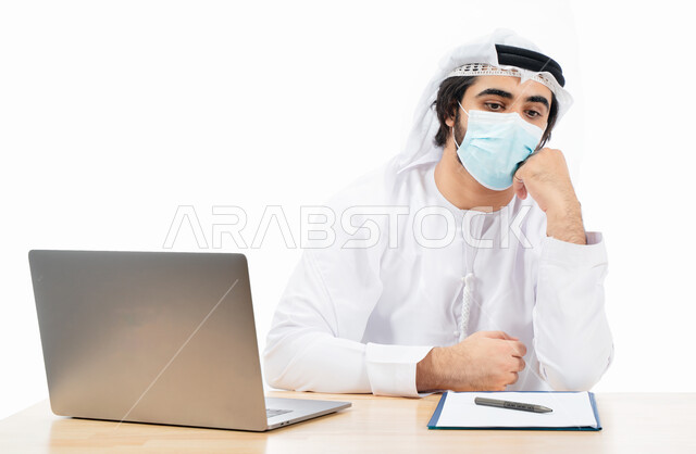 بورتريه لرجل أعمال عربي خليجي اماراتي يرتدي الكمامة الواقية، انجاز مهام العمل من خلال الحاسوب المحمول، العمل عن بعد، بيئة العمل الاماراتي، الالتزام باجراءات الوقاية من فايروس كورونا، ايماءات وتعابير تدل على التفكير والحيرة، اعمال مكتبية، خلفية بيضاء