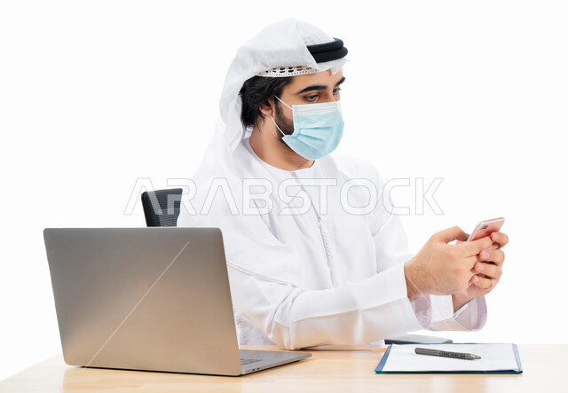 بورتريه لرجل أعمال عربي خليجي اماراتي يرتدي الكمامة الواقية ويستخدم الهاتف المحمول، انجاز مهام العمل من خلال الحاسوب المحمول، العمل عن بعد، مفهوم إدارة الأعمال، بيئة عمل، الالتزام باجراءات الوقاية من فايروس كورونا، اعمال مكتبية، خلفية بيضاء
