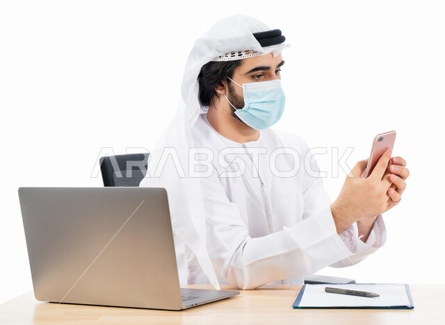 بورتريه لرجل أعمال عربي خليجي اماراتي يرتدي الكمامة الواقية ويستخدم الهاتف المحمول، انجاز مهام العمل من خلال الحاسوب المحمول، العمل عن بعد، مفهوم إدارة الأعمال، بيئة عمل، الالتزام باجراءات الوقاية من فايروس كورونا، اعمال مكتبية، خلفية بيضاء