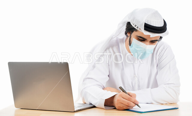 بورتريه لرجل أعمال عربي خليجي اماراتي يرتدي الكمامة الواقية، انجاز مهام العمل من خلال الحاسوب المحمول، العمل عن بعد، بيئة العمل الاماراتي، الالتزام باجراءات الوقاية من فايروس كورونا، كتابة التقارير، اعمال مكتبية، خلفية بيضاء