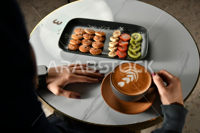 A close-up of a woman's hands holding a cup of American coffee with thick foam, a plate of small pancake pieces with fruits and chocolate, morning breakfast, snack, coffee dessert, hot cappuccino drink, delicious hot drinks