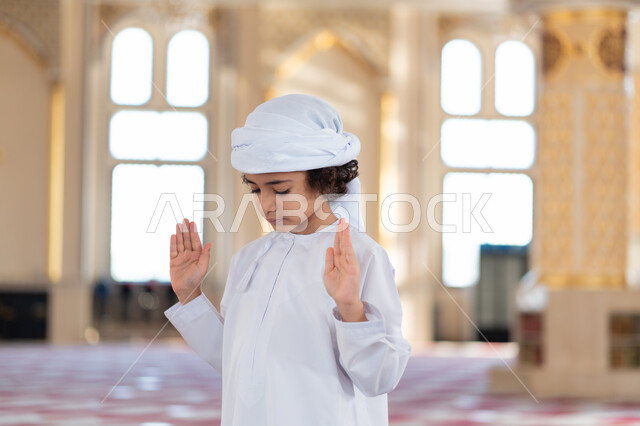 A young Arab, Gulf, Emirati boy in an Arab mosque, performing the obligatory prayer, raising his hands to enlarge the state of ihram and starting the prayer, worship and drawing closer to God, Islamic holy places, the concept of the pillars of Islam, Isla