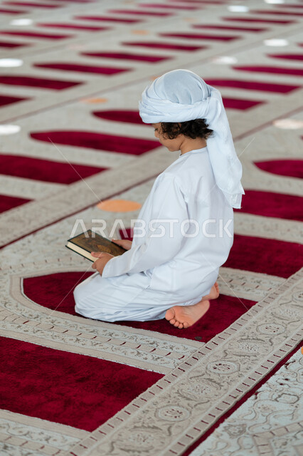 A young Arab, Gulf, Emirati boy in an Arab mosque, holding in his hand the book of the Holy Qur’an, the Book of God, the Holy Qur’an, worship and closeness to God, Islamic holy places, Islamiat and worship