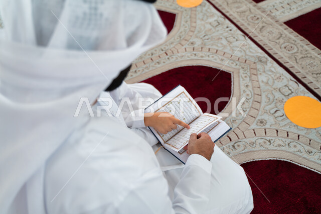 صورة مقربة لرجل عربي خليجي إماراتي وإبنه في أحد المساجد العربية، يحمل بيده المصحف الشريف ، قراءة القرآن الكريم ، تلاوة القرآن الكريم ، تحفيظ القرآن الكريم للأطفال ، العبادة والتقرب الى الله ، أماكن إسلامية مقدسة ، إسلاميات وعبادة