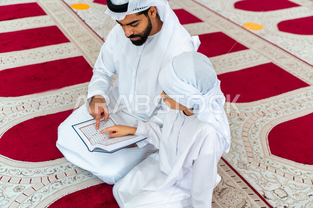 An Arab Gulf Emirati man and his son in an Arab mosque, holding the Holy Qur’an in his hand, reading the Holy Qur’an, reciting the Holy Qur’an, memorizing the Holy Qur’an for children, worship and drawing closer to God, Islamic holy places, Islamic and wo