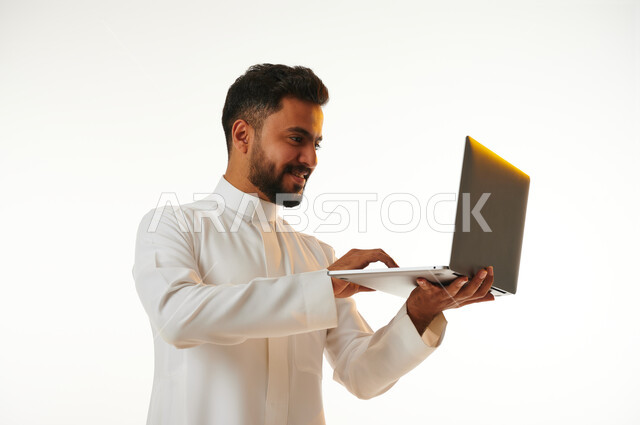 بورتريه لرجل عربي خليجي سعودي يحمل بيده جهاز الحاسوب المحمول، العمل عن بعد، انجاز مهام العمل، تصفح الانترنت، احتراف وظائف الحاسوب وتكنولوجيا المعلومات، خلفية بيضاء