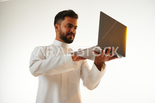 بورتريه لرجل عربي خليجي سعودي يحمل بيده جهاز الحاسوب المحمول، العمل عن بعد، انجاز مهام العمل، تصفح الانترنت، احتراف وظائف الحاسوب وتكنولوجيا المعلومات، خلفية بيضاء