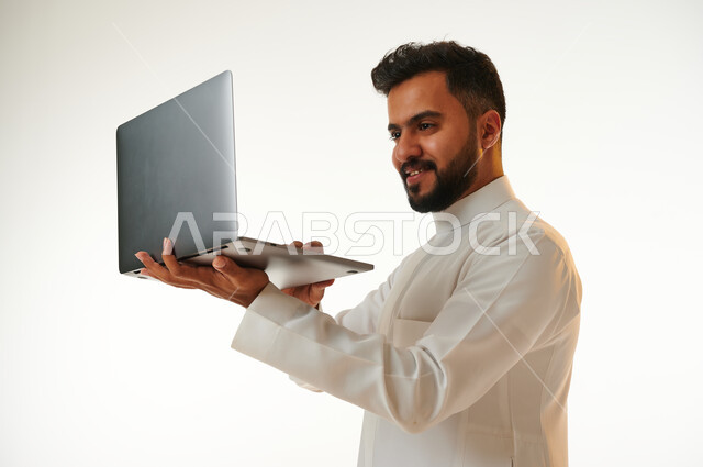 بورتريه لرجل عربي خليجي سعودي يحمل بيده جهاز الحاسوب المحمول، العمل عن بعد، انجاز مهام العمل، تصفح الانترنت، احتراف وظائف الحاسوب وتكنولوجيا المعلومات، خلفية بيضاء