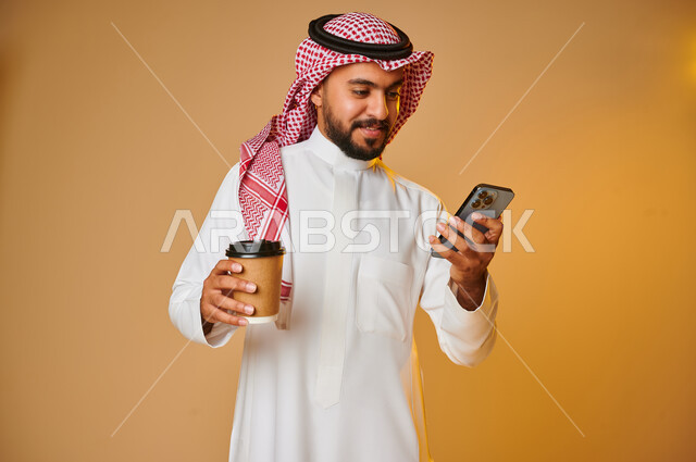 Portrait of a Saudi Arab Gulf youth with a cup in his hand and using a mobile phone, enjoying hot drinks, online shopping, online requests, smart shopping, online purchase, express delivery services for hot drinks, browsing websites, colored background
