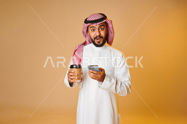 بورتريه لشاب عربي خليجي سعودي بيده كوب ويستخدم الهاتف المحمول، الاستمتاع بالمشروبات ساخنة، التسوق الالكتروني، طلبات اونلاين، التسوق الذكي، عملية شراء الكترونية، خدمات التوصيل السريع للمشروبات الساخنة، ايماءات وجه تدل على الدهشة والتعجب، خلفية ملونة