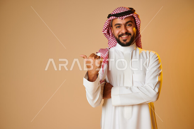بورتريه لرجل عربي خليجي سعودي يشير باصبع الابهام نحو شيء ما، ايماءات وجه مبتسمة، السعادة والسرور، حركة اصبع واشارات، خلفية ملونة