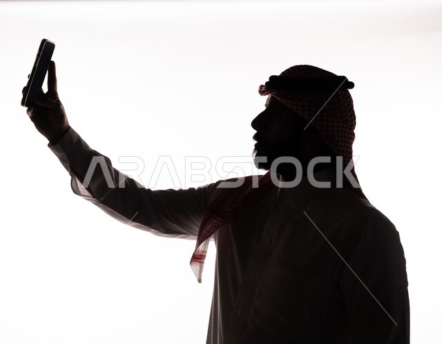 بورتريه ، صورة ظلية لرجل عربي خليجي سعودي ، إلتقاط صور سيلفي تذكاريه بإستخدام الهاتف المحمول ، إستخدام اجهزة تقنية حديثة ، التواصل الإجتماعي ، بث مباشر ، خلفية بيضاء