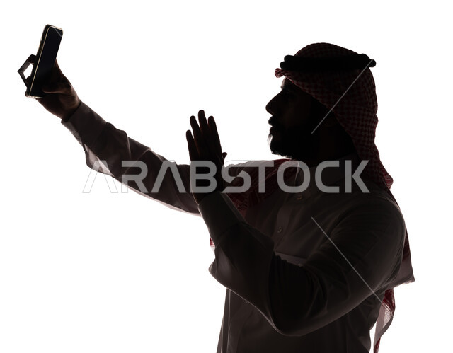 بورتريه ، صورة ظلية لرجل عربي خليجي سعودي ، إجراء مكالمة فيديو بإستخدام الهاتف المحمول ، إستخدام اجهزة تقنية حديثة ، الإتصال والدردشة ، التواصل الإجتماعي ، بث مباشر ، خلفية بيضاء