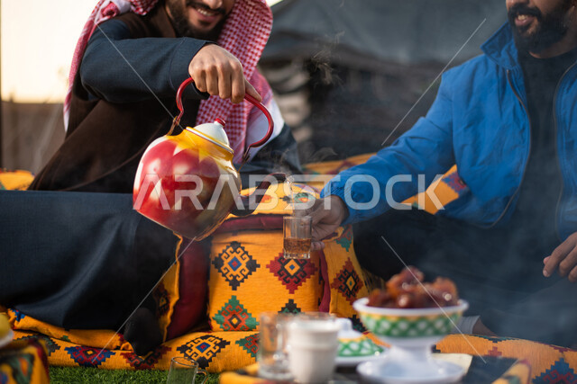 Close-up pictures of two Saudi Arabian Gulf youths enjoying a wild camping trip, wearing warm winter clothes, serving hot tea to guests, hospitality and reception, friendship and acquaintance, a popular Arab righteousness session, popular tents in the des