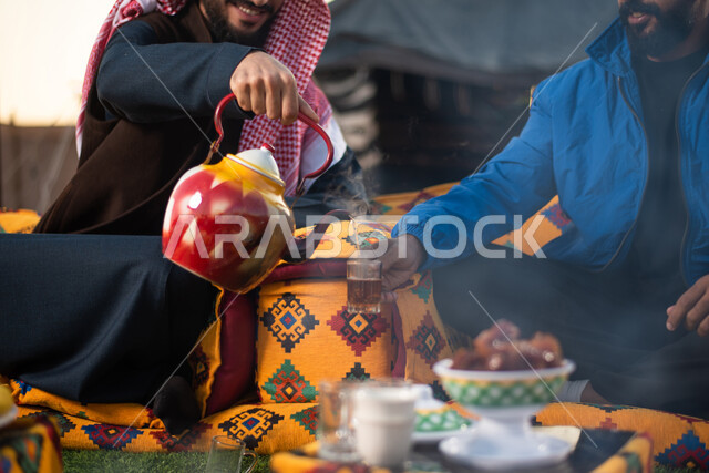 صور مقربة لشابان عربيان خليجيان سعوديان يستمتعان برحلة تخييم برية، ارتداء ملابس الشتاء الدافئة، تقديم الشاي الساخن للضيوف، حسن الضيافة والإستقبال، صداقة وتعارف، جلسة بر عربية شعبية، خيام شعبية في الصحراء، كشتة بر شبابية