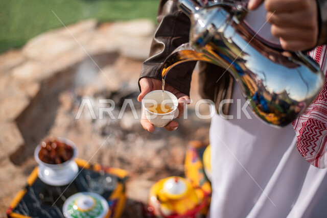 رجل عربي خليجي سعودي يقوم بصب القهوة العربية، دلة قهوة عربية، تقديم القهوة الساخنة للضيوف، رحلة تخييم برية في الصحراء، حسن الضيافة والإستقبال، جلسة بر عربية شعبية، مخيمات سياحية في صحاري السعودية، كشتة بر شبابية