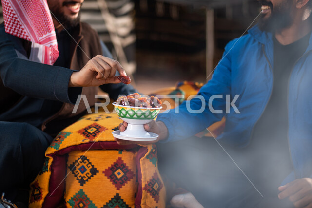 صورة مقربة لصديقان عربيان خليجيان سعوديان يستمتعان برحلة تخييم برية في الصحراء، تقديم التمر للضيوف، حسن الضيافة والإستقبال، جلسة بر عربية شعبية، خيام شعبية في صحاري السعودية، كشتة بر شبابية