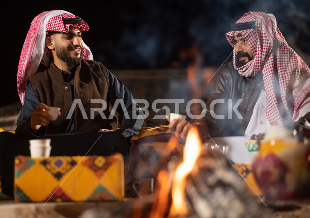 A close-up of a flame of fire, two Saudi Arabian Gulf men dressed in warm winter clothes and enjoying a coffee, a wild camping trip, a popular Arab land session, tourist camps in the deserts of Saudi Arabia, an evening youth campfire, a winter night atmos