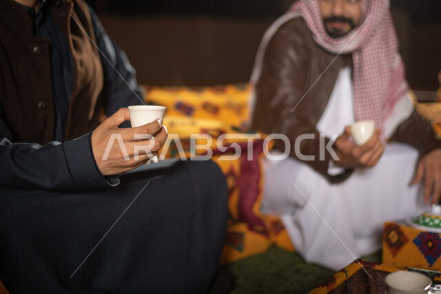 صورة مقربة لرجلان عربيان خليجيان سعوديان يرتديان ملابس الشتاء الدافئة ويستمتعان بشرب القهوة، رحلة تخييم برية، جلسة بر عربية شعبية، مخيمات سياحية في صحاري السعودية، كشتة بر شبابية مسائية، اجواء شتوية ليلية