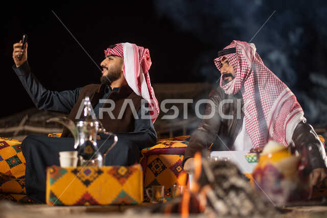 رجلان عربيان خليجيان سعوديان يرتديان ملابس الشتاء الدافئة، التقاط صورة تذكارية من خلال الهاتف المحمول، اجراء مكالمة فيديو، رحلة تخييم برية، جلسة بر عربية شعبية مع شعلة الحطب، مخيمات سياحية في صحاري السعودية، كشتة بر شبابية مسائية، اجواء شتوية ليلية