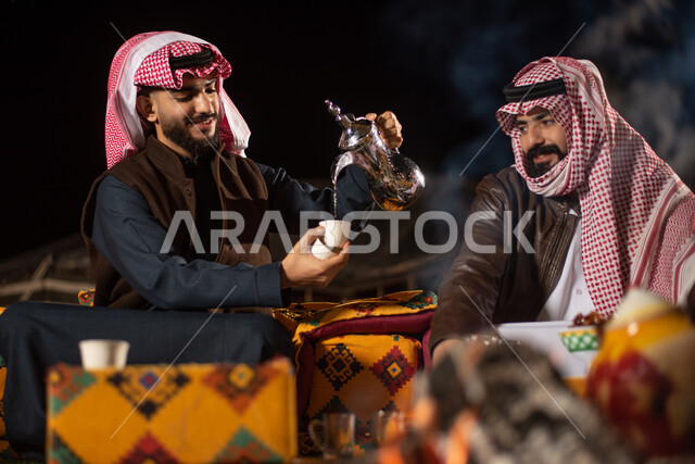 رجلان عربيان خليجيان سعوديان يرتديان ملابس الشتاء الدافئة، جلسة بر عربية شعبية مع شعلة النار، رحلة تخييم برية، تقديم القهوة الساخنة للضيوف، حسن الضيافة والاستقبال، مخيمات سياحية في صحاري السعودية، كشتة بر شبابية مسائية، اجواء شتوية ليلية