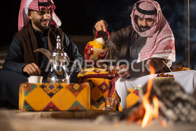 Two Saudi Arab Gulf men dressed in warm winter clothes, serving hot tea to guests, a popular Arab session with a firewood torch, good hospitality and reception, tourist camps in the deserts of Saudi Arabia, an evening youth burial, a winter night atmosphe