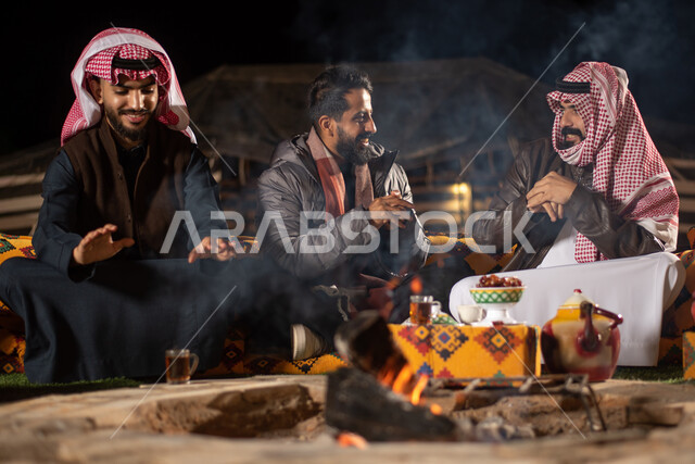 مجموعة من الاصدقاء العرب الخليجيين السعوديين يرتدون ملابس الشتاء الدافئة بايماءات تدل على البرد الشديد، التدفئة من خلال شعلة النار، حوارات ونقاشات بين الاصدقاء، خيام شعبية في صحاري السعودية، كشتة بر شبابية مسائية، اجواء شتوية ليلية