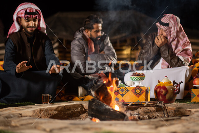 مجموعة من الاصدقاء العرب الخليجيين السعوديين يرتدون ملابس الشتاء الدافئة بايماءات تدل على البرد الشديد، التدفئة من خلال شعلة النار، حوارات ونقاشات بين الاصدقاء، خيام شعبية في صحاري السعودية، كشتة بر شبابية مسائية، اجواء شتوية ليلية