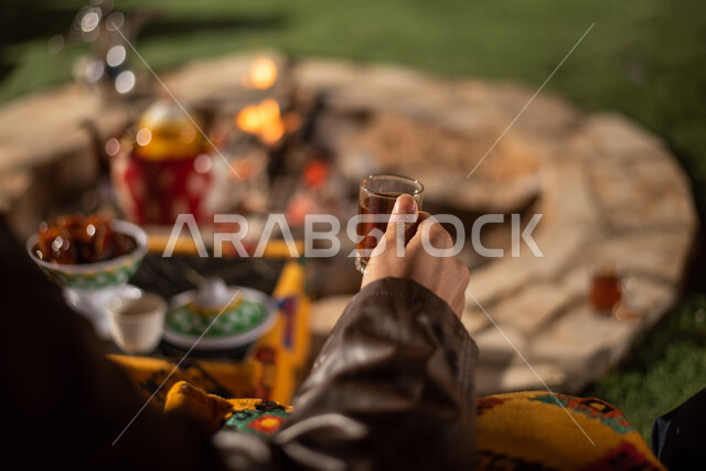 A close-up of a Saudi Arab Gulf man sitting in front of a firewood torch and enjoying drinking hot tea, a popular Arab session, a wild camping trip in the desert, popular tourist camps in the deserts of Saudi Arabia, an evening youth campfire, a winter ni