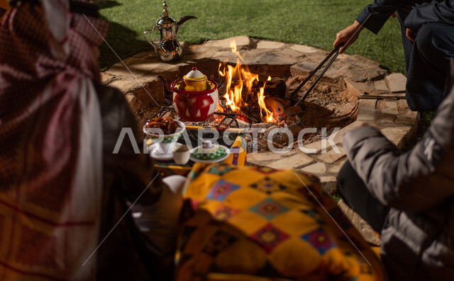 صورة مقربة لمجموعة من الاصدقاء العرب الخليجيين السعوديين يجلسون امام شعلة الحطب، التدفئة من خلال شعلة النار، الاستمتاع بالمشروبات الساخنة، مخيمات سياحية شعبية في صحاري السعودية، كشتة بر شبابية مسائية، اجواء شتوية ليلية