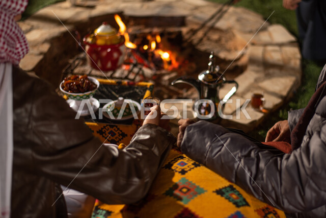 صورة مقربة لمجموعة من الاصدقاء العرب الخليجيين السعوديين يجلسون امام شعلة الحطب، التدفئة من خلال شعلة النار، الاستمتاع بالمشروبات الساخنة، مخيمات سياحية شعبية في صحاري السعودية، كشتة بر شبابية مسائية، اجواء شتوية ليلية